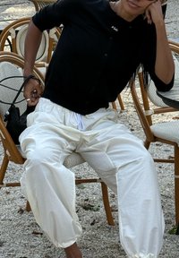 Άτομο που φοράει μαύρο πουκάμισο με κουμπιά και λευκό φαρδύ παντελόνι κάθεται σε καρέκλα σε υπαίθριο καφέ, ακουμπώντας το κεφάλι στο χέρι.