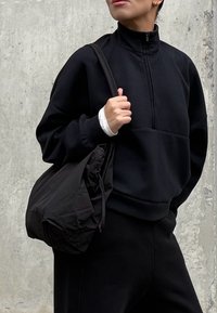 Sudadera negra con cremallera y cuello alto, bolsillo frontal, acompañada de un bolso tote negro. Textura suave y corte relajado.