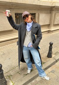 Γυναίκα με σκούρο μακρύ παλτό, ανοιχτό μπλε τζιν, καπέλο μπέιζμπολ, γυαλιά ηλίου, κρατώντας μια κούπα καφέ, στέκεται σε πεζοδρόμιο της πόλης.