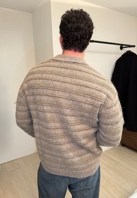 Homme aux cheveux bouclés portant un pull beige à rayures vertes et un jean bleu, debout à l'intérieur avec un manteau noir accroché sur un porte-manteau à proximité.