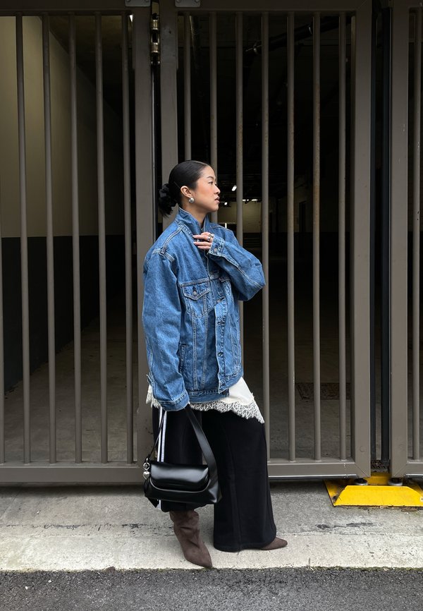 Femeie într-o jachetă denim supradimensionată, pantaloni largi negri, cizme maro, ținând o geantă neagră, stând în profil în fața unor porți înalte de metal.