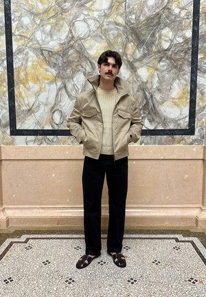 Chaqueta beige con dos bolsillos en el pecho, suéter de punto color crema, jeans oscuros y sandalias marrones. Se encuentra sobre un azulejo estampado frente a un fondo texturizado.