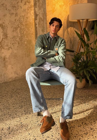 Chaqueta verde con botones amarillos sobre una camisa de rayas, combinada con jeans azul claro y zapatos marrones con cordones naranjas, sentado en una silla.