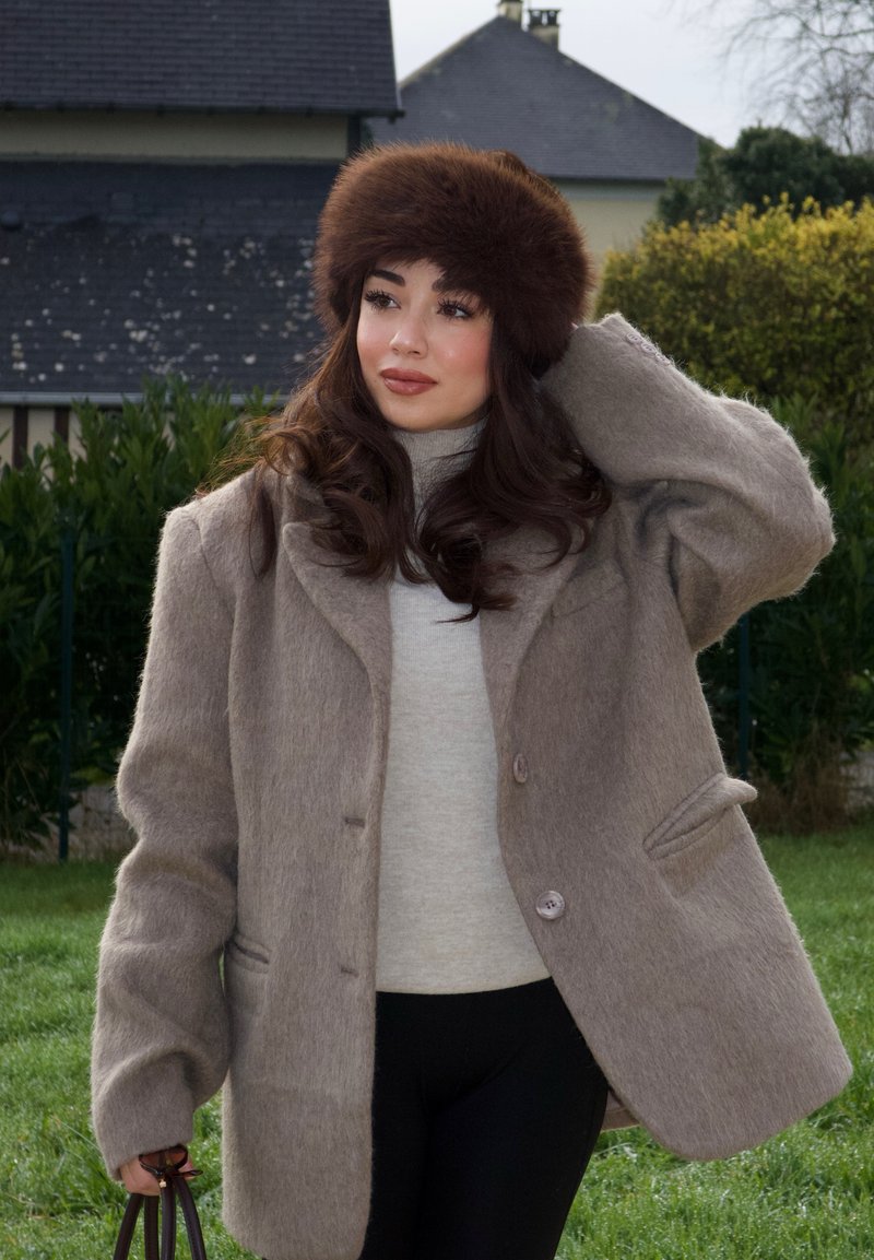 Brown wool-blend oversized coat with large buttons, paired with a light gray turtleneck. A dark fur hat adds texture. Black pants complete the outfit.