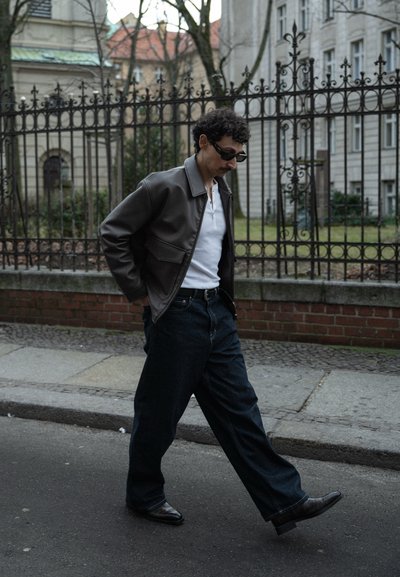 Chaqueta de cuero marrón sobre una camisa blanca, combinada con unos vaqueros de pierna ancha azul oscuro y elegantes zapatos negros, caminando en un entorno urbano.