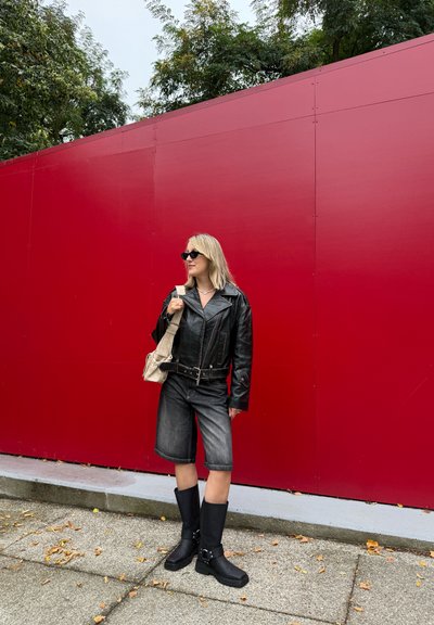 Chaqueta de cuero negra con un gran cuello, pantalones cortos de mezclilla y botas negras hasta la rodilla, de pie contra una pared roja. Se muestra un bolso de hombro casual.