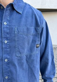 Camisa azul de botões feita de tecido suave, com um bolso no peito, detalhes cosidos e um rótulo da marca no bolso.