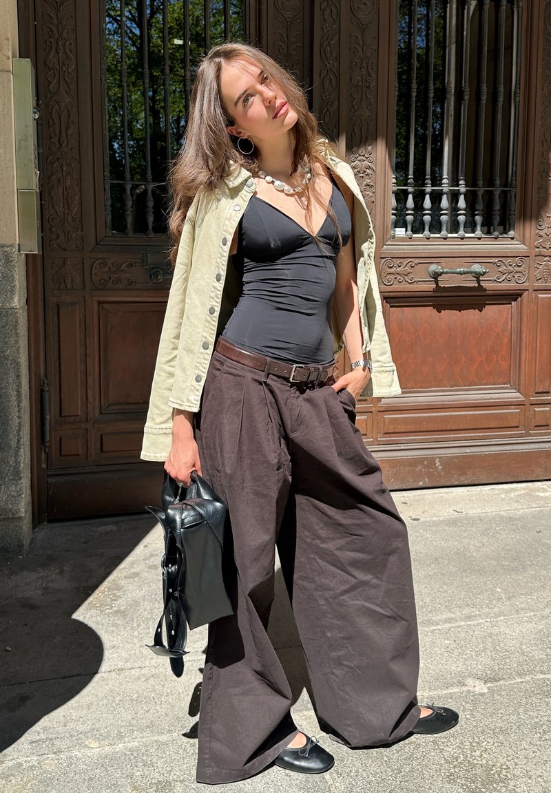 Jeune femme portant une veste beige sur un haut noir, pantalon large marron, ballerines noires, tenant un sac noir, debout près de portes en bois ornées.