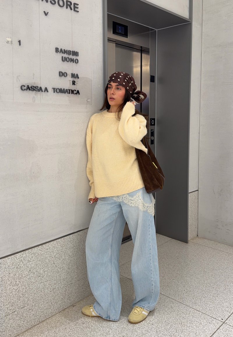 Maglione lavorato a maglia color crema, jeans a gamba larga azzurri chiaro con dettagli in pizzo, borsa marrone e foulard marrone a pois, in piedi vicino a un ascensore.