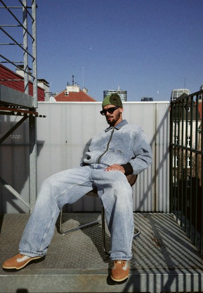 Conjunto de denim con una chaqueta de cremallera en la parte delantera y pantalones de corte holgado en azul claro, complementado con un gorro verde y gafas de sol negras. Zapatos marrones.