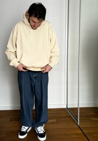 Krém színű kapucnis pulóver, kenguruzsebbel, laza kék farmernadrággal és fekete-fehér sportcipőkkel párosítva. A háttérben fa padló és tükör látható.