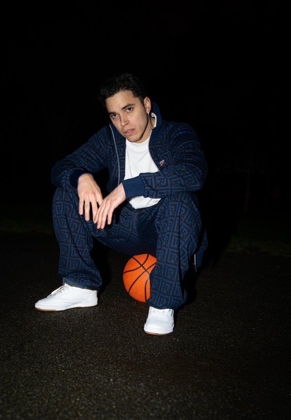 Man hurkt buiten 's nachts en rust op een oranje basketbal, draagt een blauw patroon trainingspak, wit t-shirt en witte sneakers.