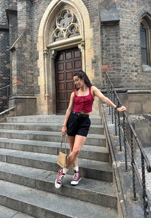 Top corto rojo, pantalones cortos negros de cintura alta, zapatillas burdeos altas con calcetines blancos y un pequeño bolso beis, posando en unas escaleras de piedra.