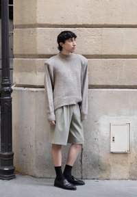 Beige grovstrikket genser, lys grønn plissert shorts, svarte sokker og loafers. Står mot en teksturert steinvegg.