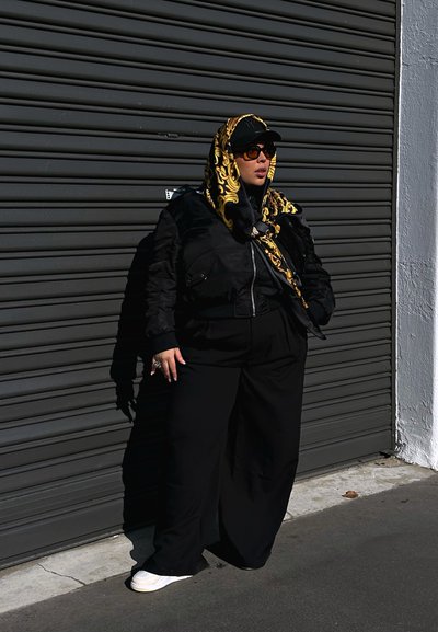 Chaqueta bomber negra con cremallera, pantalones anchos negros y zapatillas blancas. También son visibles una bufanda con un patrón dorado, gafas de sol y una gorra.