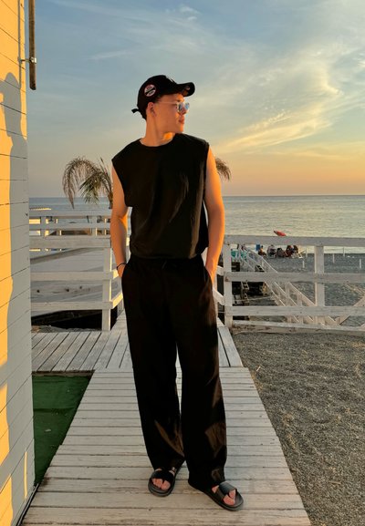 Camiseta negra sin mangas, pantalones negros de corte relajado y sandalias negras. Fondo con un brillante atardecer, plataforma de madera y palmeras visibles.