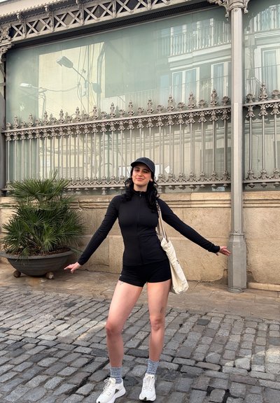 Top negro de manga larga con cremallera y pantalones cortos negros, combinados con zapatillas blancas y una gorra oscura. Lleva un bolso tote beige colgado del hombro.