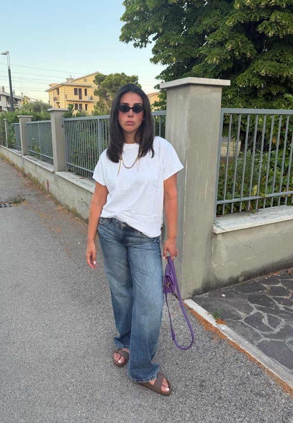 Kvinde iført solbriller, hvid t-shirt, vide jeans og sandaler står på fortovet og holder en lilla håndtaske nær et metalhegn og træer.