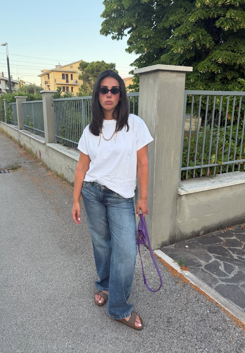 Femme portant des lunettes de soleil, t-shirt blanc, jean large et sandales, debout sur le trottoir tenant un sac à main violet près d'une clôture en métal et des arbres.