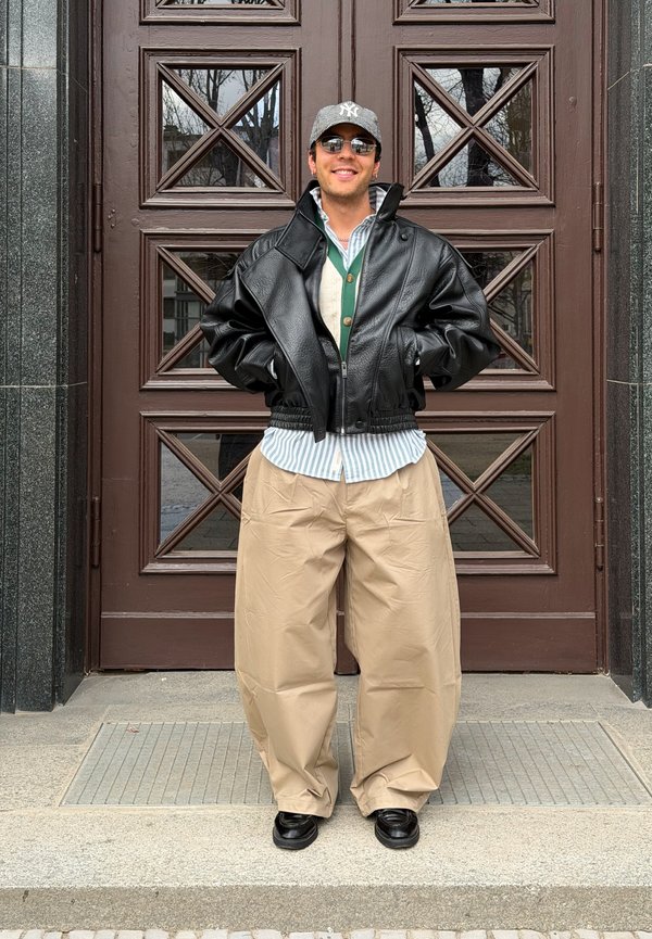 Tânăr purtând o șapcă gri, o jachetă neagră de piele, o cămașă cu dungi, un pulover verde, pantaloni largi bej și pantofi negri, stând în fața unei uși de lemn.