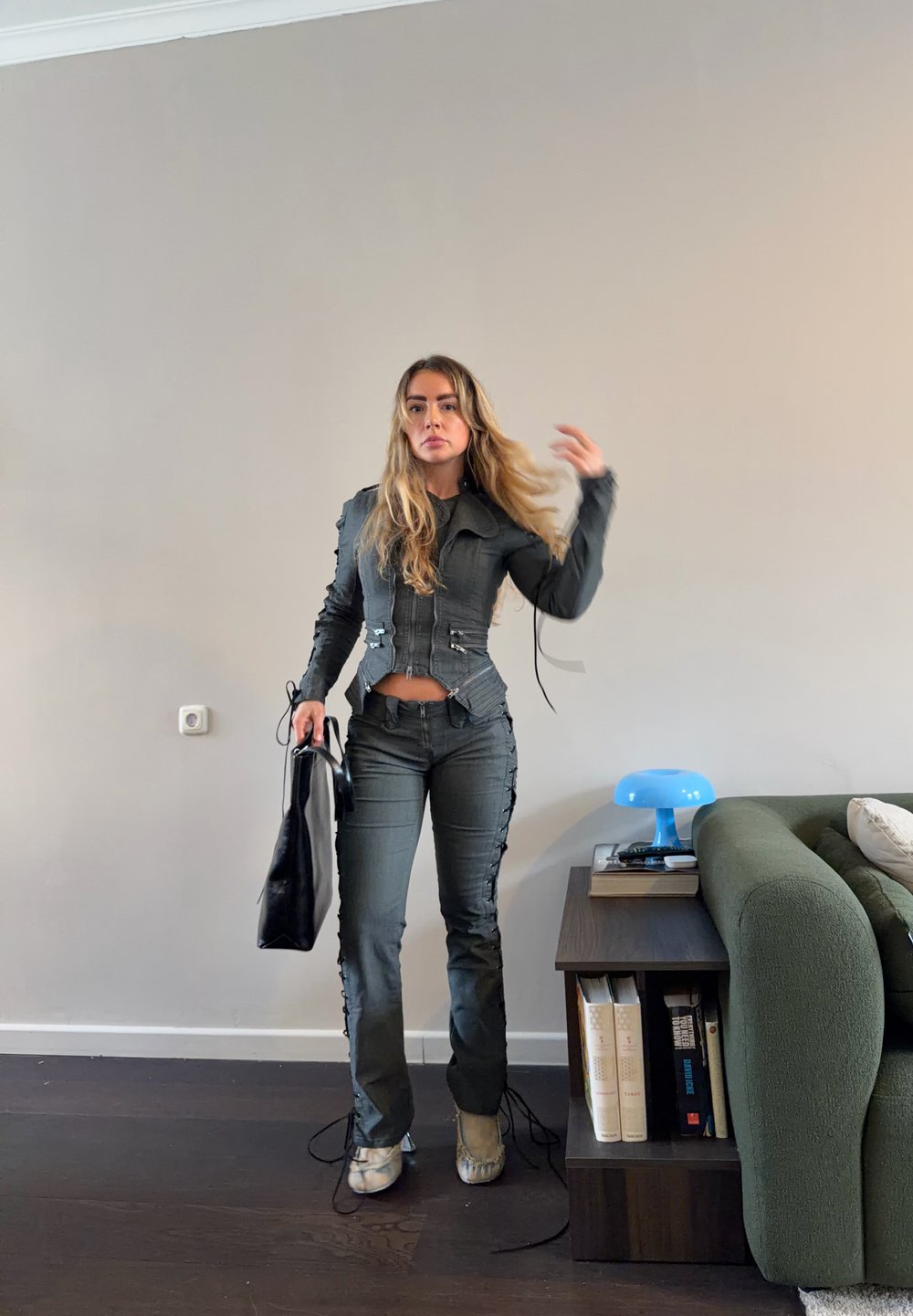 Femme aux longs cheveux portant une veste cintrée sombre et un pantalon, tenant un sac noir, debout dans le salon près d'un canapé vert et d'une table d'appoint avec des livres et une lampe bleue.