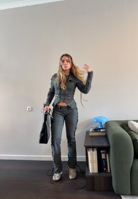 Femme aux longs cheveux portant une veste cintrée sombre et un pantalon, tenant un sac noir, debout dans le salon près d'un canapé vert et d'une table d'appoint avec des livres et une lampe bleue.