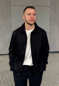 Man med kort hår och skägg som bär en svart jacka över en vit skjorta, stående mot en grå kaklad vägg med händerna i fickorna.