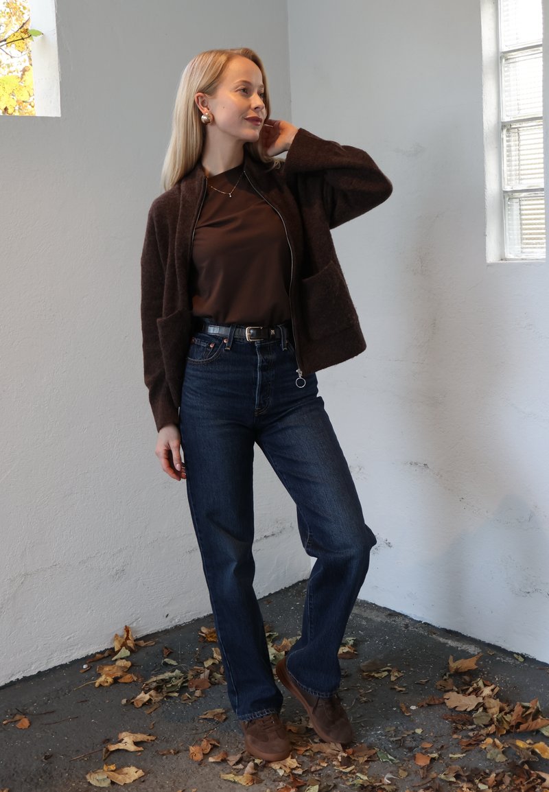 Brun cardigan med lynlås, brun top, højtaljede mørke denimjeans og brune sko. Modellen står mod en hvid væg med efterårsblade nedenfor.