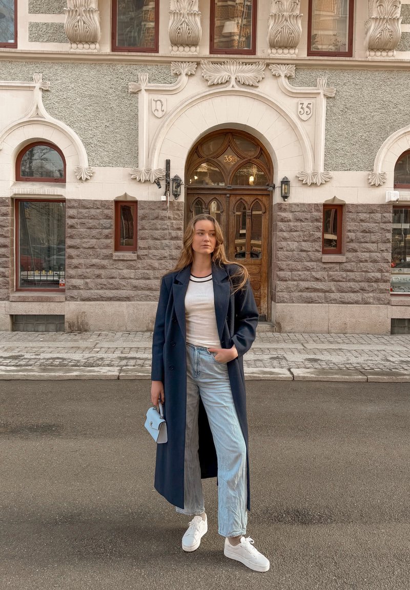 Giovane donna con cappotto lungo blu navy, camicia bianca, jeans chiari e sneakers bianche si trova in piedi sulla strada davanti a un edificio in pietra ornato con porta in legno.