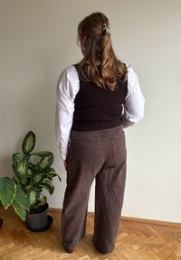 Brown corduroy wide-leg trousers paired with a black sleeveless top over a white long-sleeve shirt. The background includes a green houseplant.
