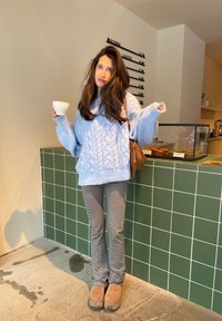 Femme aux longs cheveux bruns portant un pull bleu torsadé et un pantalon rayé tient une tasse blanche à l'intérieur d'un café derrière un comptoir en carrelage vert.