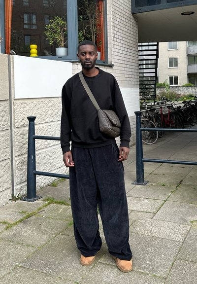 Sudadera negra oversize, pantalones de terciopelo negro holgados, pantuflas marrones y un bolso cruzado marrón. De pie en un área al aire libre pavimentada.