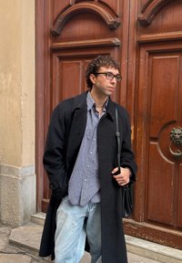 Homme aux cheveux bouclés et lunettes, portant un manteau noir, une chemise à carreaux, un jean clair, et portant un sac à bandoulière noir, debout près d'une grande porte en bois.