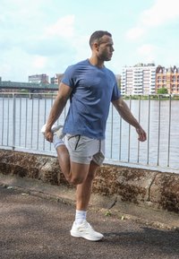 Zils sporta t-krekls ar teksturētu rakstu, gaiši pelēkas šortus, baltas sporta apavus, kas stiepjoties atrodas pie ūdeņiem ar pilsētas ainu fonā.