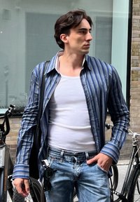 Modellen har på seg en blå stripete skjorte med knapper over en hvit tanktopp, sammen med lyse denimjeans. Sorte solbriller holdes i hånden.