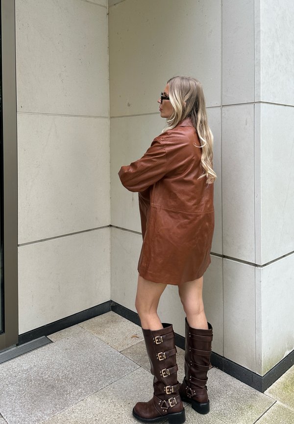 Veste en cuir marron oversized, bottes marron montantes avec plusieurs accents de boucles, devant un mur en pierre de couleur claire.
