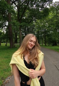 Zwarte top met ruches, gecombineerd met een gele gebreide trui over de schouders. Lange, golvende blonde haren en een groene parkachtergrond.