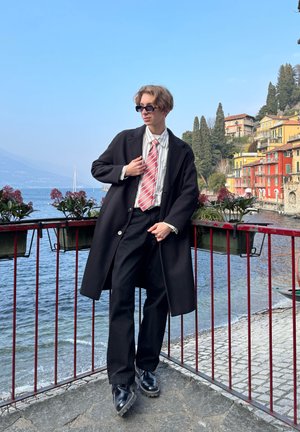 Abrigo negro, camisa blanca, corbata de rayas rojas, pantalones negros y botas negras brillantes. De pie cerca de una vista al agua con edificios coloridos de fondo.