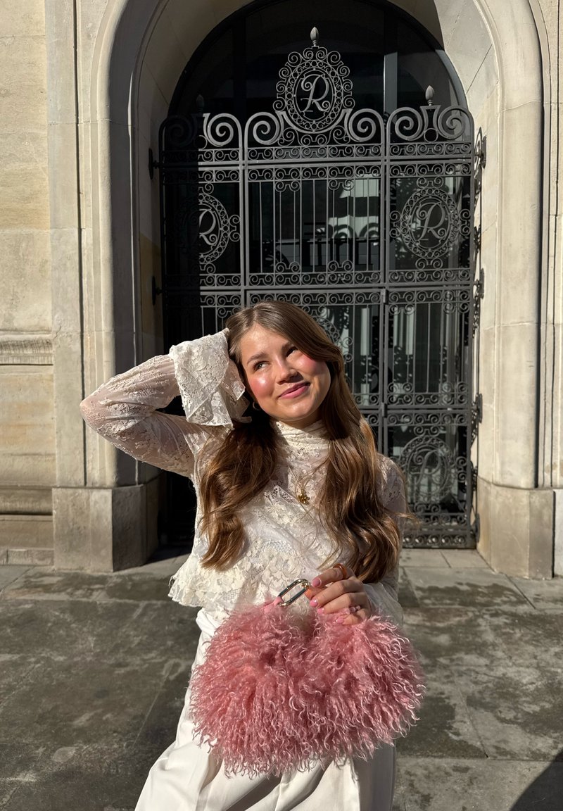 Jeune femme en blouse blanche en dentelle tenant un sac rose duveteux, posant avec un sourire devant une porte en métal noir ornée et une arche en pierre.