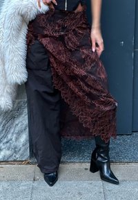 Monikerroksinen viininpunainen pitsihame mustien housujen päällä, jossa on monimutkaiset kukkakuvioinnit. Mustat, kärjellään terävät kengät täydentävät asun.