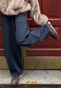 Veste en fausse fourrure beige, pantalon large noir, et baskets sombres avec des touches de rose. La personne est posée avec une jambe levée, devant une porte rouge.