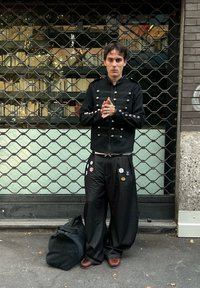 Veste noire avec des clous en métal, pantalon noir ample orné de divers badges, chaussures foncées et un sac de sport noir. Arrière-plan avec une grille métallique.
