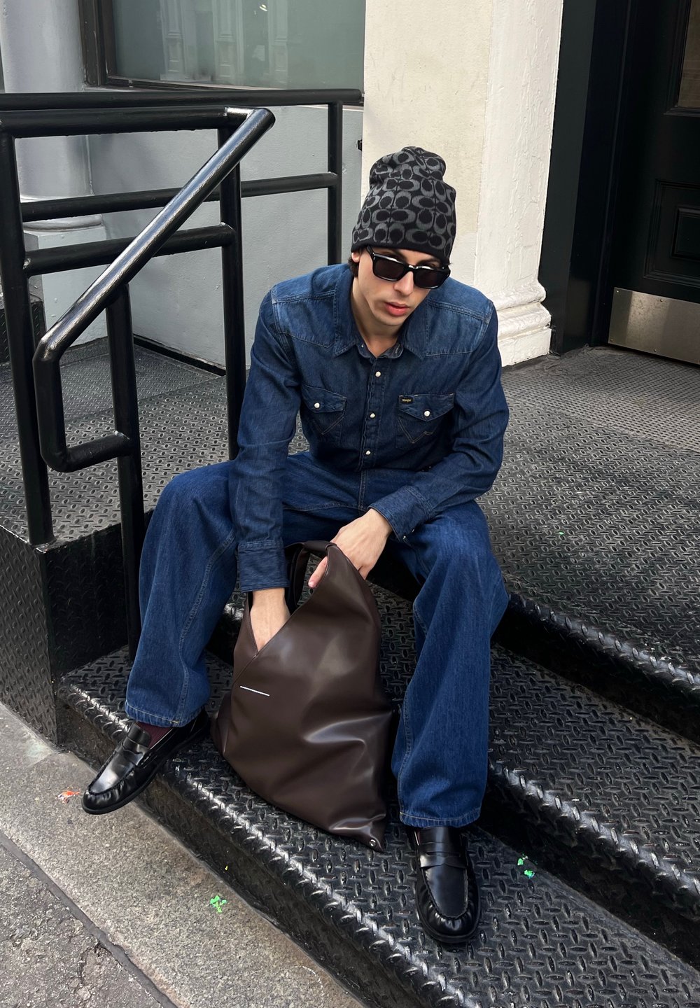 Camisa de ganga, jeans de perna larga, óculos de sol pretos e um gorro estampado. Asegura uma mala de mão em couro castanho com uma forma distinta.