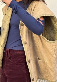 Egy kék, hosszú ujjú felsőt viselő személy, amelynek a vállai kivágottak, bordó kordbársony nadrágot és egy világosbézs steppelt mellényt visel, nagy zsebekkel.