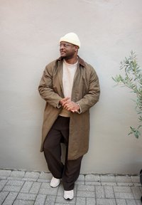 Parka maro cu guler maro, pulover bej, pantaloni maro în carouri și adidași crem; accesorizat cu o căciulă galbenă și ochelari.