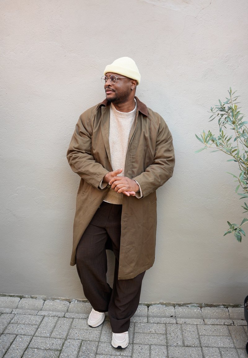 Parka marron avec un col marron, pull beige, pantalon marron à carreaux, et baskets crème ; accessoirisés d'un bonnet jaune et de lunettes.