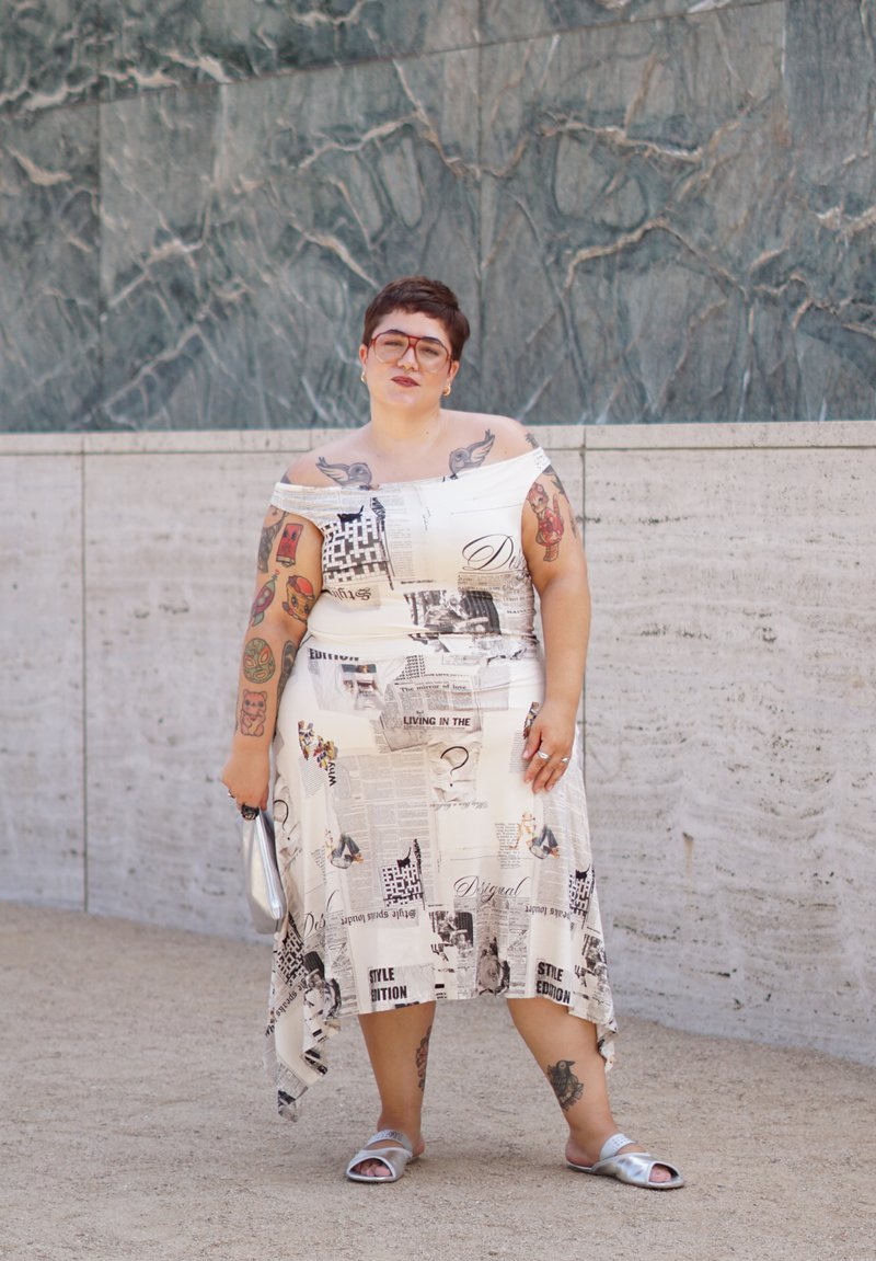 Personne aux cheveux courts et portant des lunettes, vêtue d’une robe imprimée journal à épaules dénudées et de sandales argentées, debout devant un mur de pierre texturé.