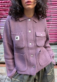 Femme portant un cardigan en tricot mauve avec quatre poches avant et des boutons argentés, se tenant devant un fond de volets rose et gris.