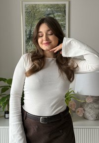 Hvit ribbestrikket genser med lange ermer og utsvingte mansjetter, kombinert med et brunt skjørt og svart belte. Innendørs setting med plante og lampe i bakgrunnen.