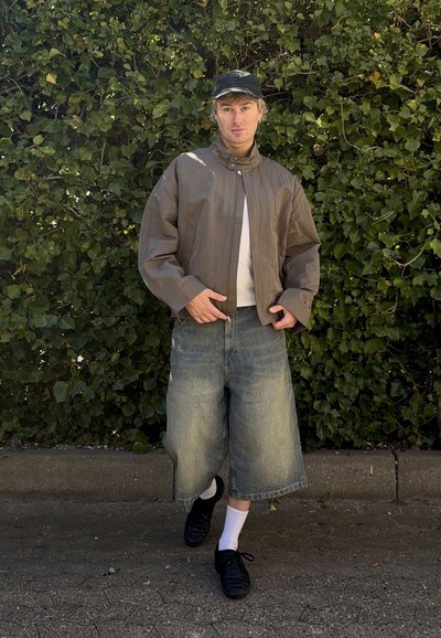 Joven con chaqueta corta marrón, pantalones cortos anchos de mezclilla, gorra negra, calcetines blancos y zapatos negros, de pie al aire libre frente a un fondo frondoso.